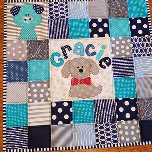May include: A blue and grey patchwork quilt with a white centre panel featuring the name "Gracie" in blue letters and a grey dog with a red bow tie.