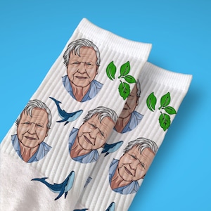 May include: White socks with a repeating pattern of a man's face, a whale, and a green leaf. The man's face is a cartoon illustration of a person with brown hair and a white beard.