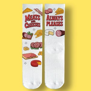 May include: Pair of white socks with a fun design of meats and cheeses. The left sock has the text "Meats and Cheeses" in red, and the right sock says "Always Pleases." Illustrations of cheese, salami, and bacon are scattered across the socks.