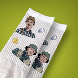 May include: White ribbed socks with illustrations of two men, a gold coin, and two silver can tabs. One man wears a brown cap and headphones, the other a green hat and headphones. The background is a lime green colour.