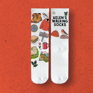 May include: White socks featuring illustrations of walking-related items such as boots, a compass, a sandwich, a sheep, a red jacket, a map, a mug, a bird, and a hat. The text "Helen's Walking Socks" is printed on the socks.