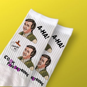 May include: Pair of white ribbed socks with illustrations of a smiling man in a green vest and the text "A-HA!". Additional text includes "COOK PASS BABTRIDGE" and a mug with "SAVOURY 99".