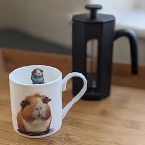 May include: White ceramic mug with a brown and white guinea pig illustration. The mug has a black handle and is sitting on a wooden surface. A black french press is in the background.