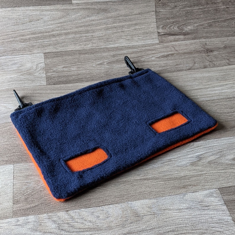 May include: A blue fleece hammock with two orange fleece openings. The hammock has two black clips on the top.