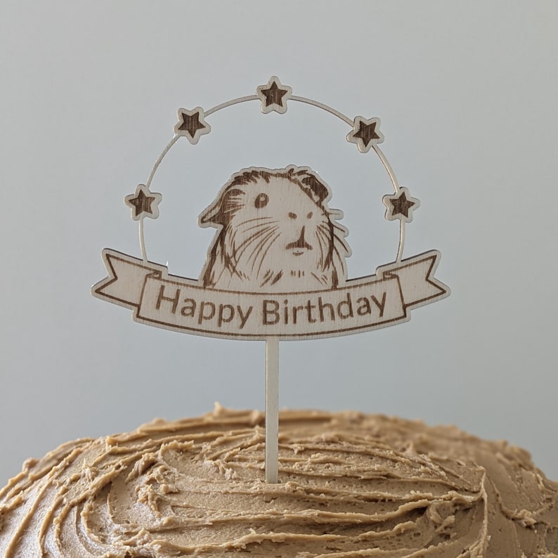 May include: Wooden cake topper with a guinea pig illustration and the text "Happy Birthday" in a banner. The topper is decorated with stars.