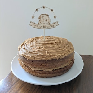 May include: A brown frosted cake with a wooden birthday cake topper featuring a guinea pig and the text "Happy Birthday".