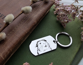 May include: A silver keychain with a black and white line drawing of a guinea pig on a rectangular tag. The keychain is on a green surface with a book and a sprig of white flowers.
