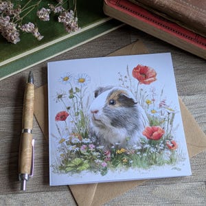 Peut inclure: Carte de vœux avec une illustration à l'aquarelle d'un cochon d'Inde au milieu de fleurs sauvages, dont des marguerites et des coquelicots rouges. La carte est sur une enveloppe marron, avec un stylo en bois et des livres anciens en arrière-plan.