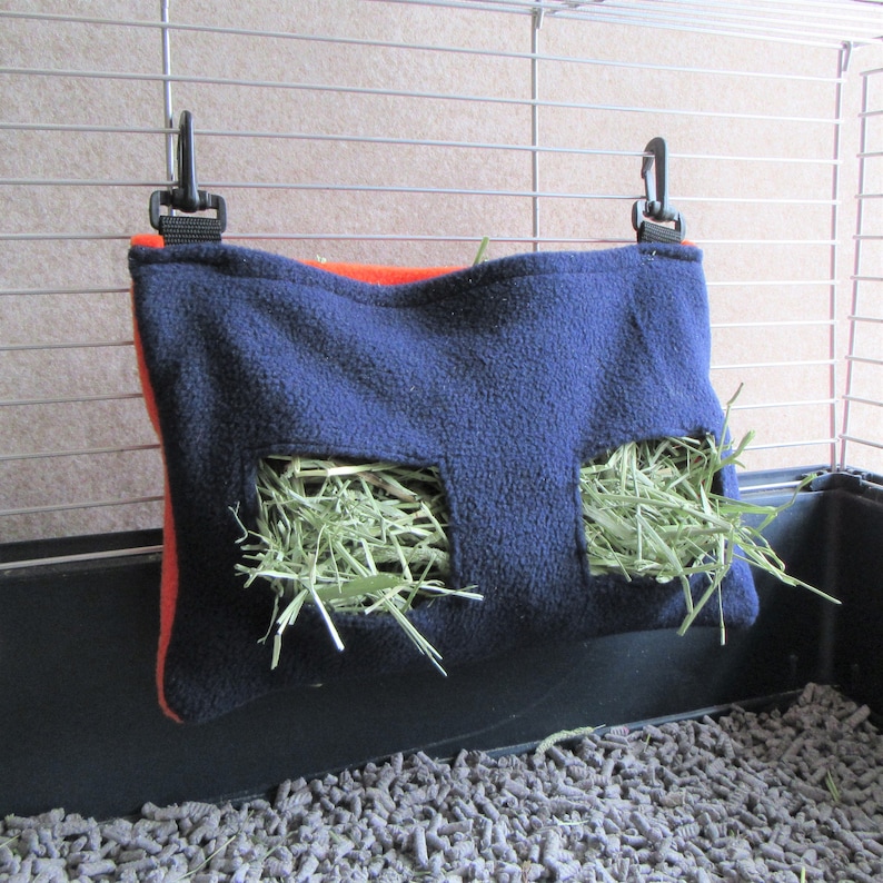 May include: A blue fleece hay feeder with two openings for hay. The feeder is hanging from a cage with orange trim and black clips.