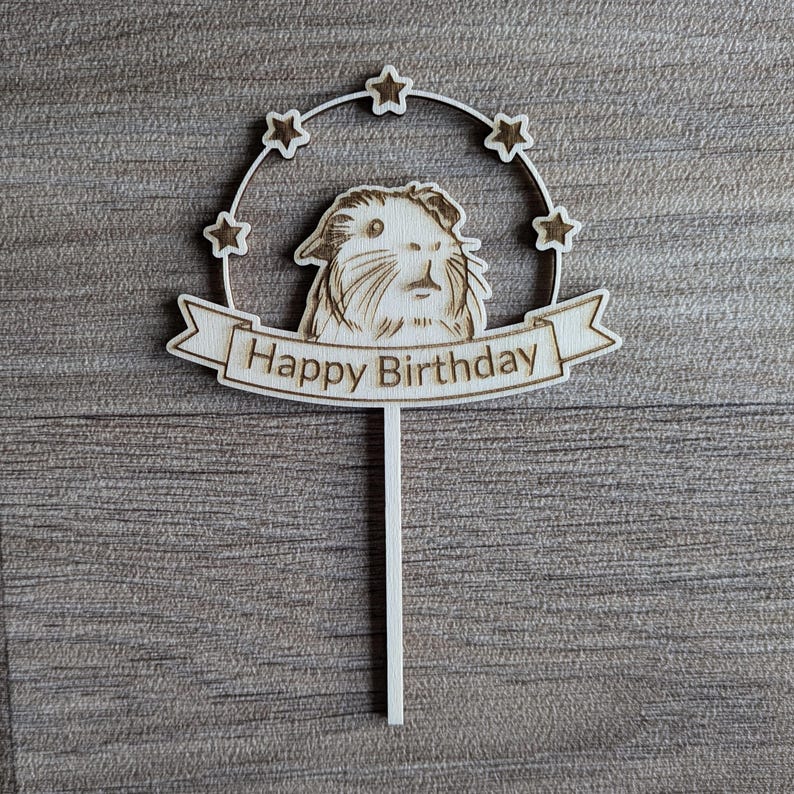 May include: Wooden cake topper with a guinea pig illustration and a banner that reads "Happy Birthday". The topper is surrounded by a circle of stars.