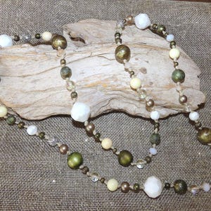 May include: Two beaded necklaces with a variety of beads in shades of white, cream, brown, and green. The necklaces are draped over a piece of driftwood, set against a neutral burlap background. The beads vary in size and shape.