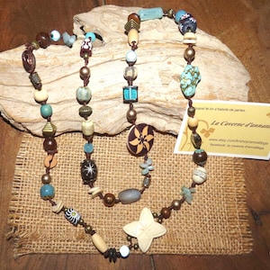 Op de afbeelding: Een handgemaakte ketting met een verscheidenheid aan kralen in bruin, beige, turquoise en blauw. De ketting bevat houten, stenen en patroon kralen, met een stervormige hanger. Het label luidt "La Caverne d'annatibga".