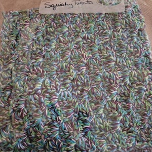 May include: A close-up of a crocheted dishcloth in a variety of colors, including green, blue, purple, and white. The dishcloth is in a plastic bag with a label that says "Squishy Twists" and "Great Value".