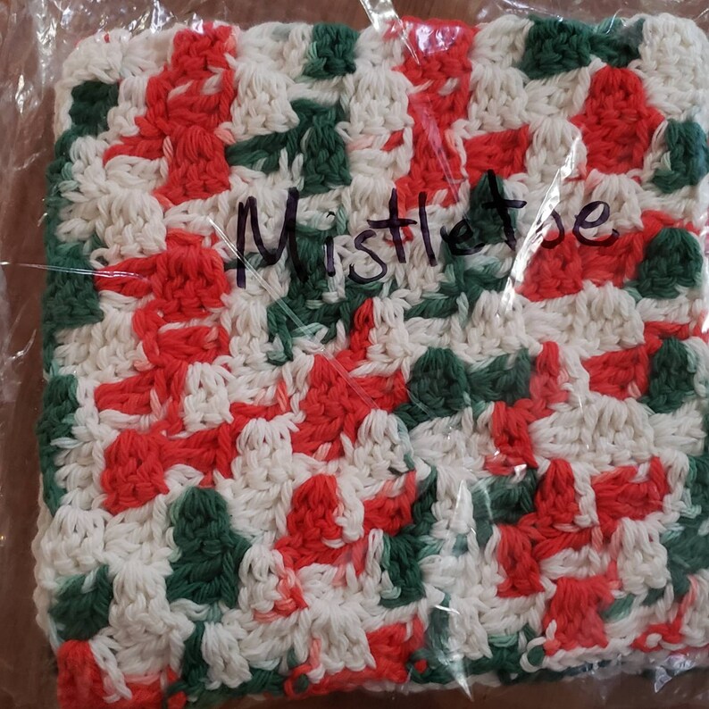 May include: A white, red, and green crocheted dishcloth with the word "Mistletoe" written on it.