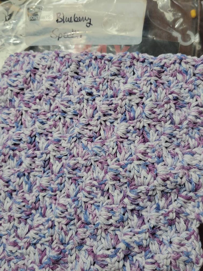 May include: A close-up of a crocheted dishcloth in shades of blue, purple, and white. The dishcloth is made with a textured, waffle-like stitch. The label reads "Blueberry Speckled".