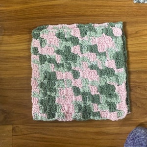 May include: A square crocheted dishcloth in pink and green. The dishcloth is made with a diagonal pattern.
