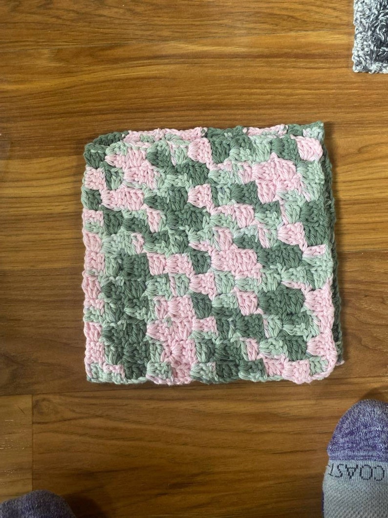 May include: A square crocheted dishcloth in pink and green. The dishcloth is made with a diagonal pattern.