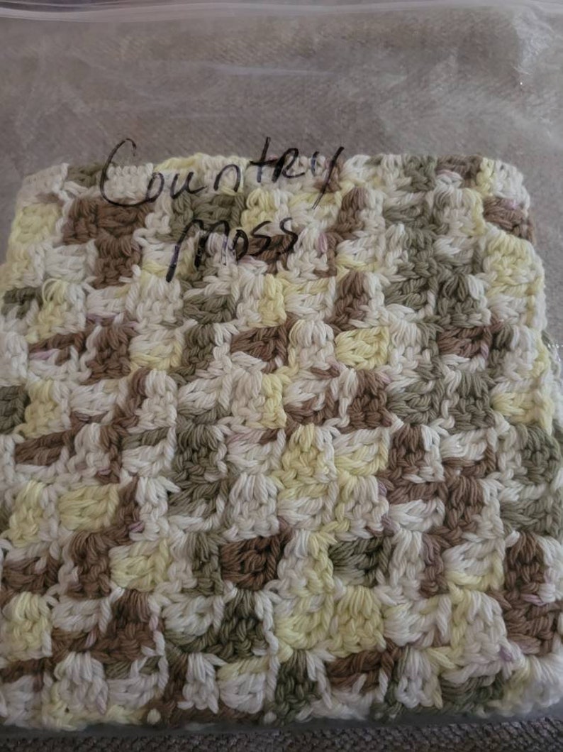 May include: A crocheted square with a country moss design in shades of white, yellow, brown, and green. The square is in a plastic bag with the words "Country Moss" written on it.