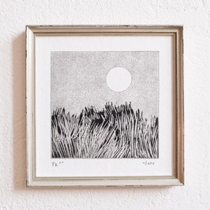 May include: A square framed print featuring a black and white ink drawing of a field of tall grass under a large circular sun. The artwork is signed PK 21 in the bottom left corner and numbered 1/100 in the bottom right corner of the white paper.