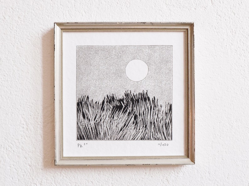 May include: A square framed print featuring a black and white ink drawing of a field of tall grass under a large circular sun. The artwork is signed PK 21 in the bottom left corner and numbered 1/100 in the bottom right corner of the white paper.