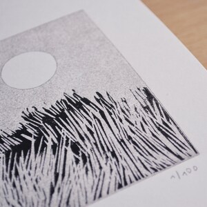 May include: A black and white linocut print on textured paper featuring a stylised landscape with tall, sharp grass blades and a solid white circular moon in a stippled grey sky. The print is numbered 1/100 in the bottom right corner.