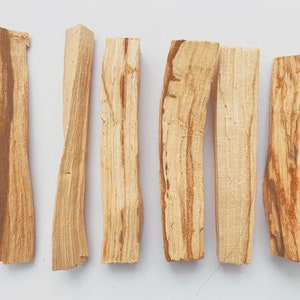 May include: A collection of six light brown palo santo wood sticks, arranged horizontally against a white background. The sticks have a natural, textured appearance with varying shades of brown and tan, showcasing the wood grain.