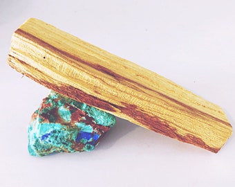 May include: A light-colored, rectangular piece of Palo Santo wood rests atop a colorful, textured stone. The wood has dark brown streaks, and the stone is a mix of blue, green, and brown hues. The items are set against a white background.