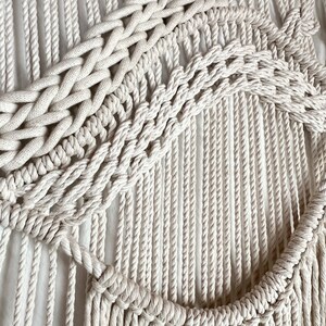 May include: A white macrame wall hanging with a geometric design. The hanging is made of thick cotton rope and has a fringe at the bottom.