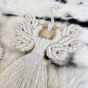 Macramé angel ornament with braided wings, a wooden bead head, and a long fringed tassel body, displayed on soft white fur