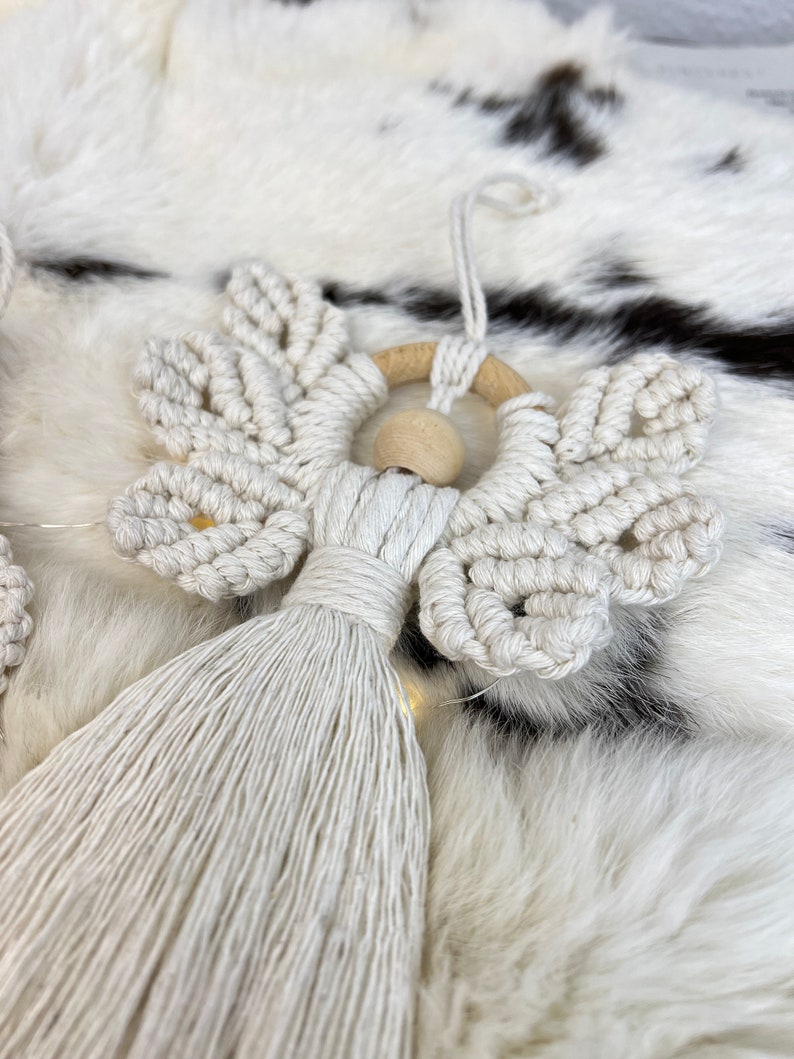 Macramé angel ornament with braided wings, a wooden bead head, and a long fringed tassel body, displayed on soft white fur
