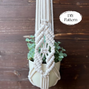 May include: Cream-colored macrame plant hanger with a woven diamond pattern, holding a white ceramic pot filled with green plants. The text "DIY Pattern" is visible in the upper right corner. The background is a dark wood.