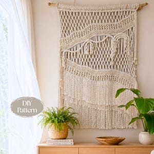 May include: A large off-white macrame wall hanging suspended from a wooden branch, featuring intricate knotted diamond patterns, horizontal woven sections, and long fringe at the bottom. The text DIY Pattern is visible in a gray oval overlay.