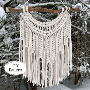 Könnte beinhalten: Cremefarbener Makramee-Wandbehang mit einem natürlichen Ast. Das handgefertigte Textil weist komplizierte Knotenmuster und lange, gefranste Stränge auf. Der Text "DIY Pattern" befindet sich in einem weißen Oval. Dekorative Boho-Wanddekoration.