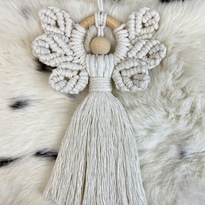 Macramé angel ornament with braided wings, a wooden bead head, and a long fringed tassel body, displayed on soft white fur