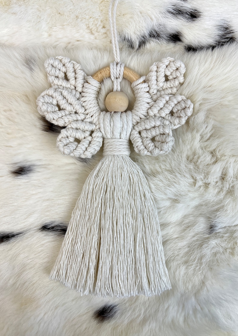 Macramé angel ornament with braided wings, a wooden bead head, and a long fringed tassel body, displayed on soft white fur
