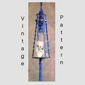 Op de afbeelding: Een blauwe macramé hanger met een witte teddybeer erin. De hanger heeft een kwast aan de onderkant.
