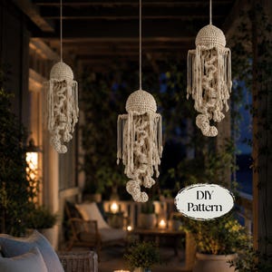 May include: Three off-white crocheted jellyfish hanging decorations suspended by strings. Each jellyfish features a rounded, textured cap and long, dangling, wavy tentacles. An oval graphic in the lower right corner displays the text DIY Pattern.