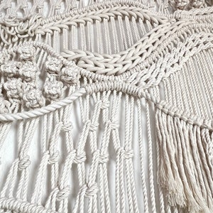 May include: A white macrame wall hanging with a variety of knots and textures. The hanging features a large tassel at the bottom.