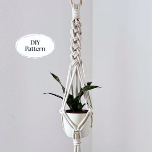 May include: A handmade macrame plant hanger in natural off-white cotton cord. The plant hanger holds a white ceramic pot with a green plant. The text "DIY Pattern" is in a white oval on the left.