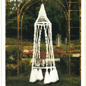 Puede incluir: Macetero colgante blanco de macramé con una plataforma redonda y borlas. El macetero está colgado de una cadena en un jardín.