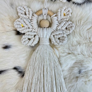 Handmade macramé angel ornament featuring textured wings, a wooden bead head, and a long tassel body, displayed on soft fur.