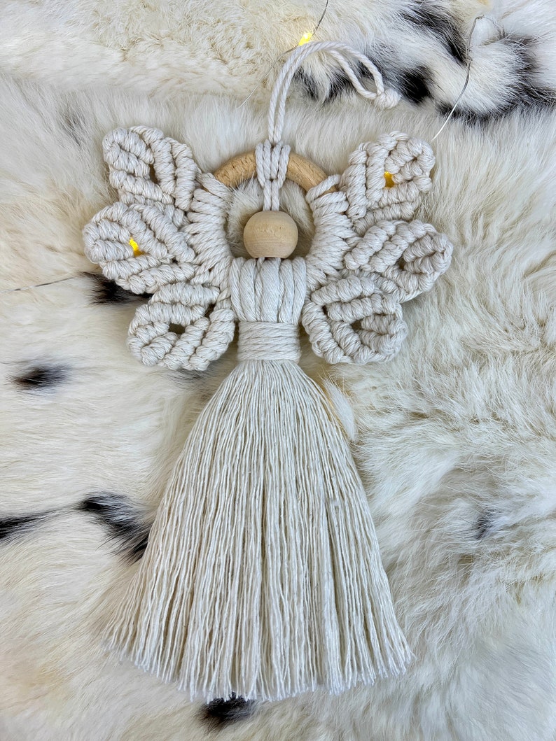 Handmade macramé angel ornament featuring textured wings, a wooden bead head, and a long tassel body, displayed on soft fur.