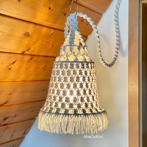 Può includere: Un paralume in macramè bianco con un bordo a frangia è appeso a un cavo bianco. Il paralume è acceso e diffonde una luce calda. Il testo "WhiteOwlKnot" è visibile nell'angolo in basso a destra dell'immagine.