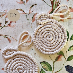 Peut inclure: Deux dessous de verre beiges, circulaires et faits à la main, avec un motif en spirale. Chaque dessous de verre a une boucle pour être suspendu. Le mot "DIY" est dans un cercle en haut à gauche. Les dessous de verre sont sur un tissu à motifs floraux.