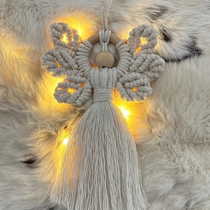Handmade macramé angel decoration with a wooden bead head and a tassel body, photographed on a fur background with fairy lights.