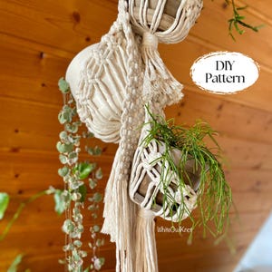 May include: Cream-colored macrame plant hanger with two wooden planters, one holding green plants. The design includes intricate knotting and fringe details. The text "DIY Pattern" is visible. The overall theme is home decor.