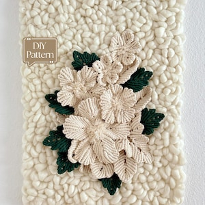 May include: A macrame wall hanging featuring a cream-colored chunky yarn background with a central arrangement of cream-colored macrame flowers and dark green macrame leaves. A brown speech bubble graphic with the text DIY Pattern is visible.