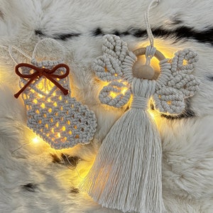 Handmade macramé Christmas ornaments — a small textured stocking with a suede bow and a tasselled angel with braided wings — displayed on soft fur with warm fairy lights behind them.