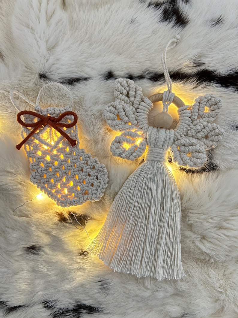 Handmade macramé Christmas ornaments — a small textured stocking with a suede bow and a tasselled angel with braided wings — displayed on soft fur with warm fairy lights behind them.