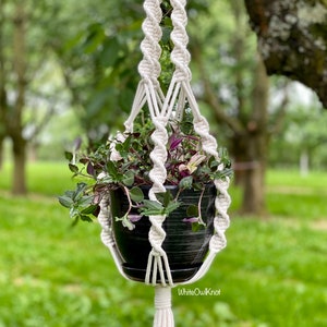 May include: A white macrame plant hanger with a black pot and green plant with purple flowers hanging from a tree branch. The hanger has a tassel at the bottom and the text "WhiteOwlKnot" is visible at the bottom of the image.